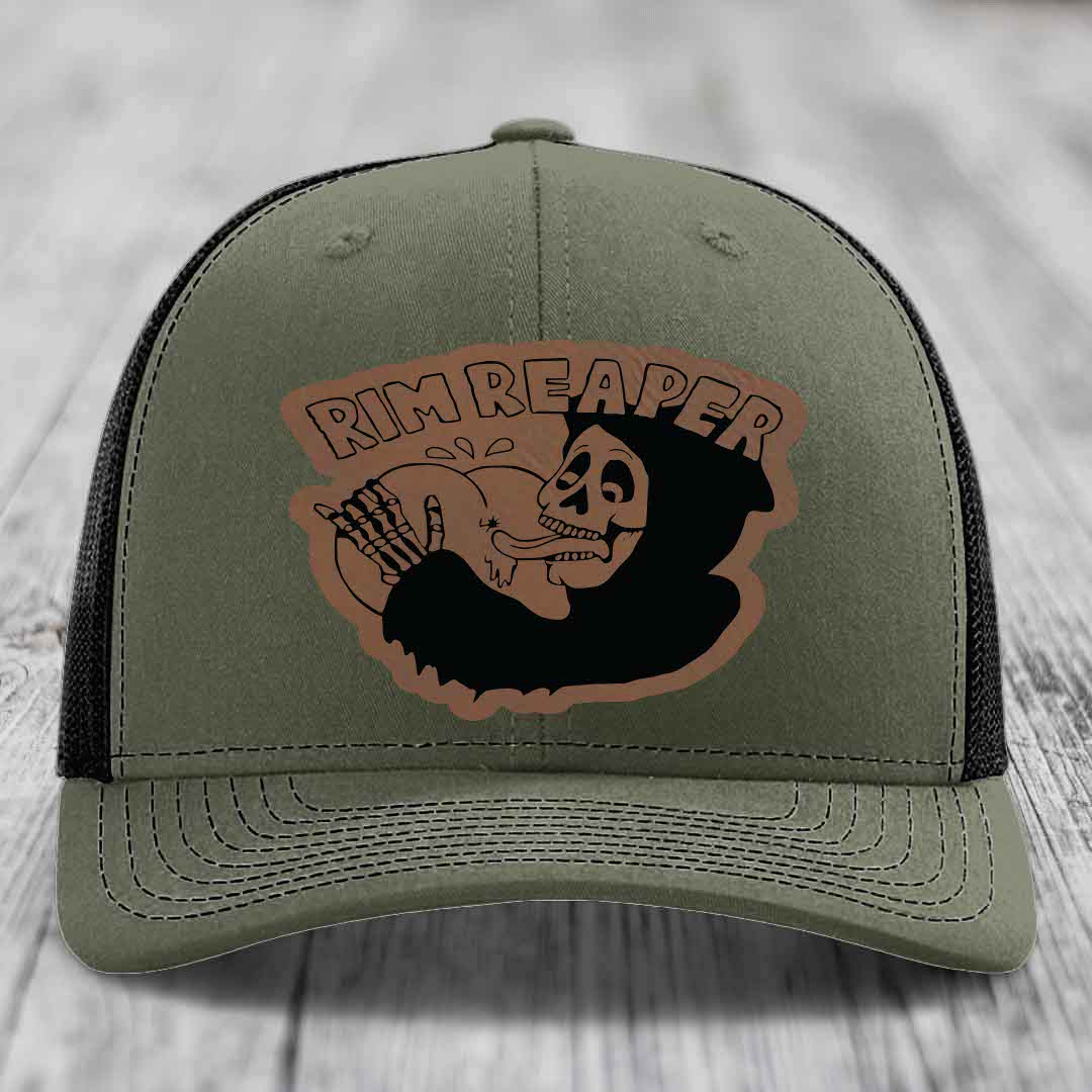 Olive green trucker hat with black mesh back and a brown leatherette patch featuring a skull holding a shotgun and the text &