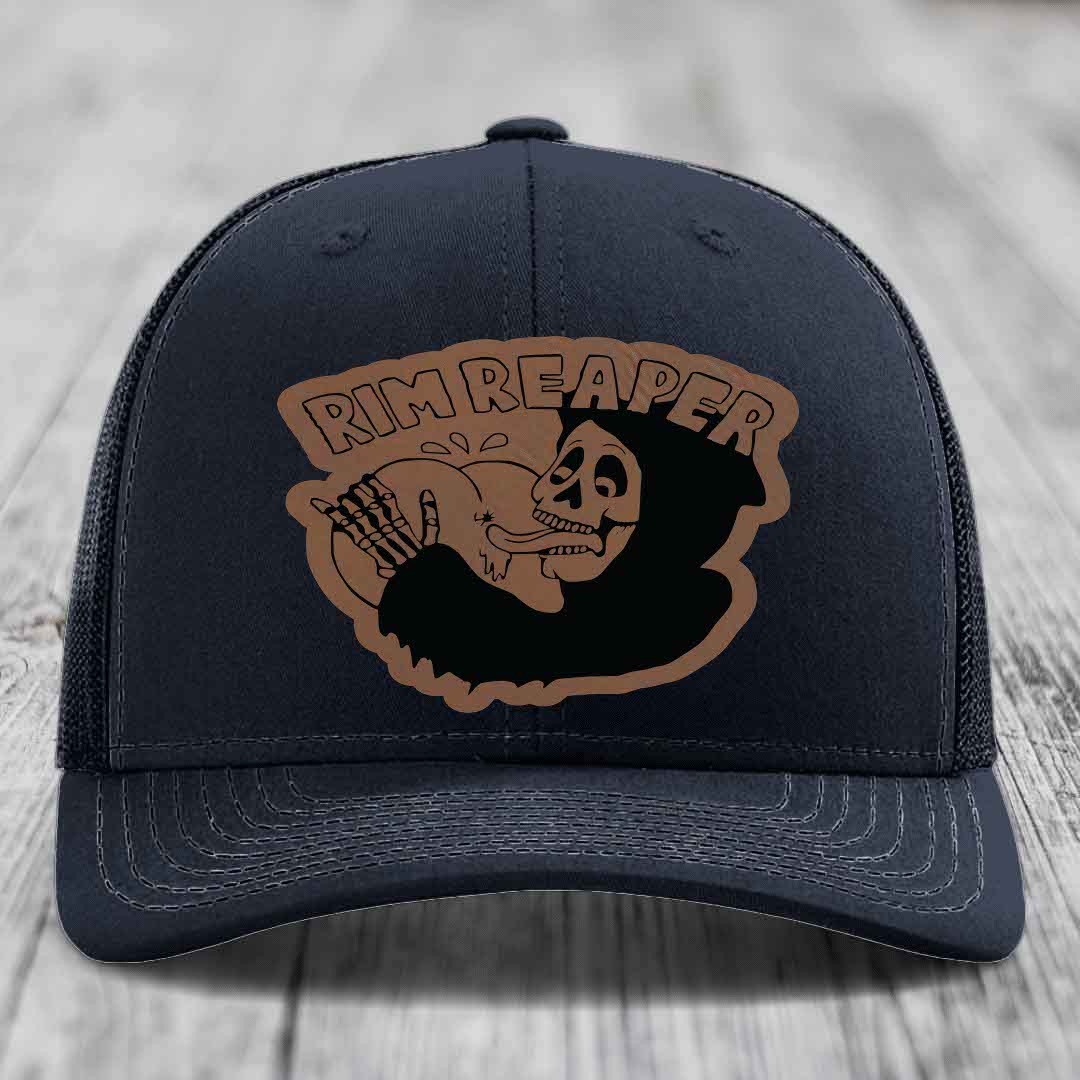 Dark blue trucker hat with brown leather patch featuring skull holding baseball, text reads &