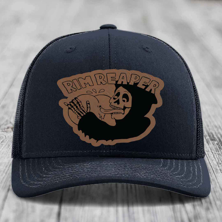 Dark blue trucker hat with brown leather patch featuring skull holding baseball, text reads &