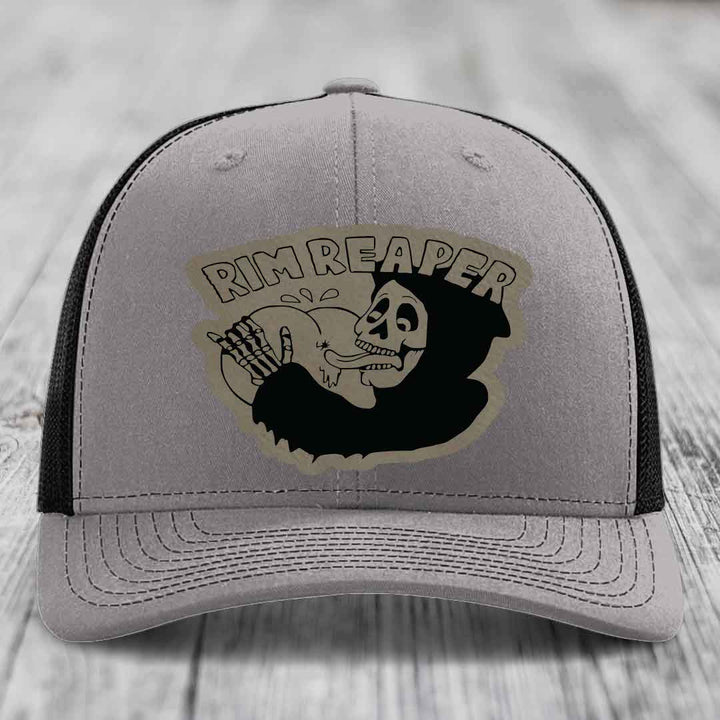 Grey trucker hat with black mesh back and a black skull-and-bones patch that says &