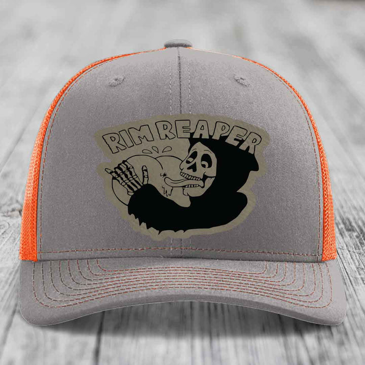 Grey trucker hat with orange mesh sides and a black skull graphic patch that says Rim Reaper.