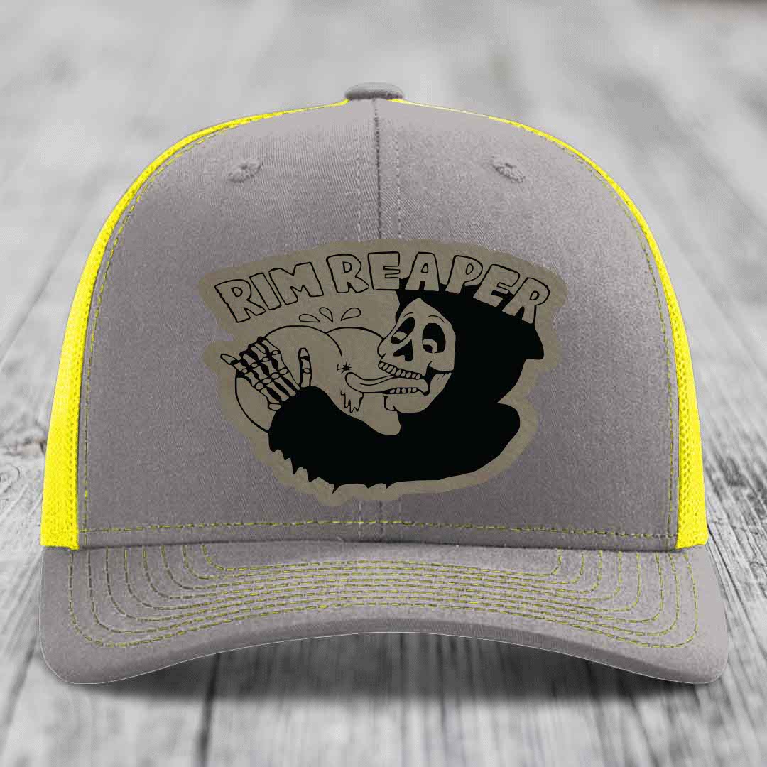 Grey trucker hat with yellow mesh and stitching, featuring a black skull graphic and &