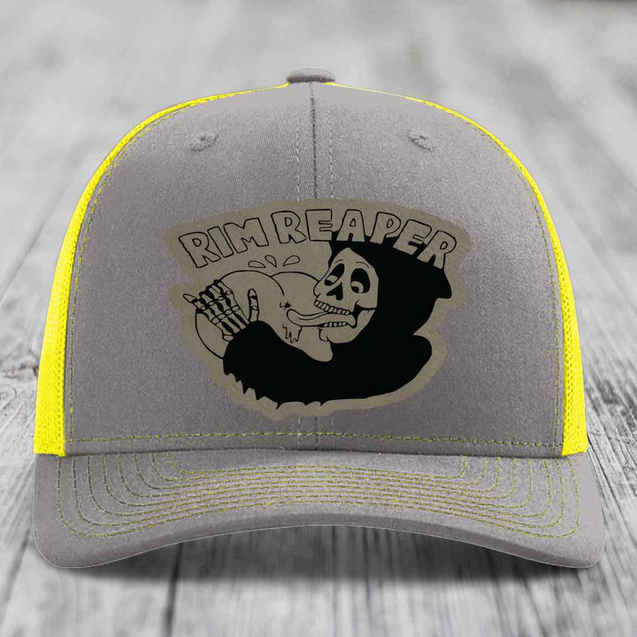 Grey trucker hat with yellow mesh and stitching, featuring a black skull graphic and &