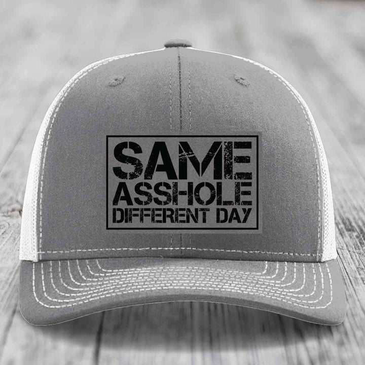 Grey trucker hat with white mesh back and black embroidered patch reading &