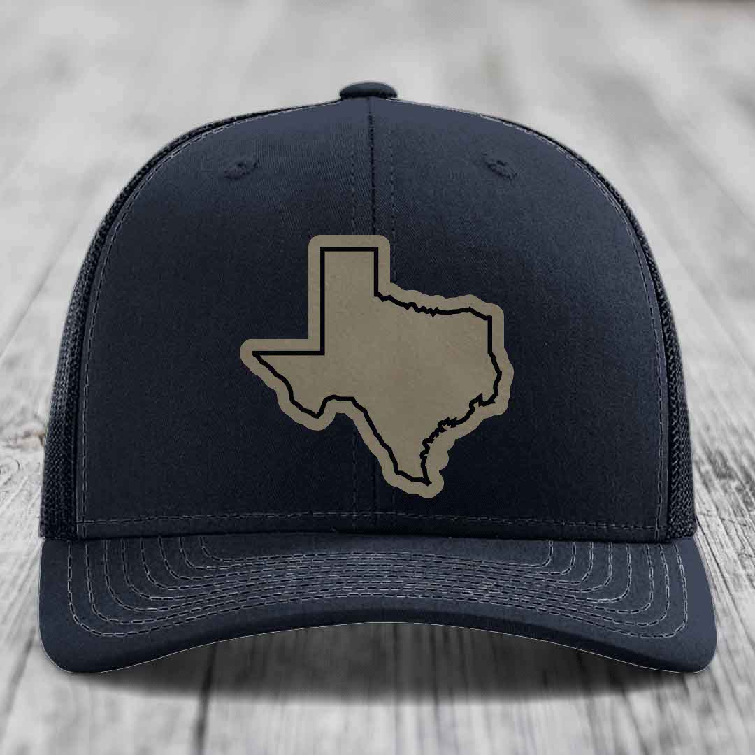 Black trucker hat with tan Texas state outline patch on front, placed on wooden surface.