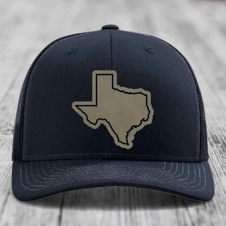 Black trucker hat with tan Texas state outline patch on front, placed on wooden surface.