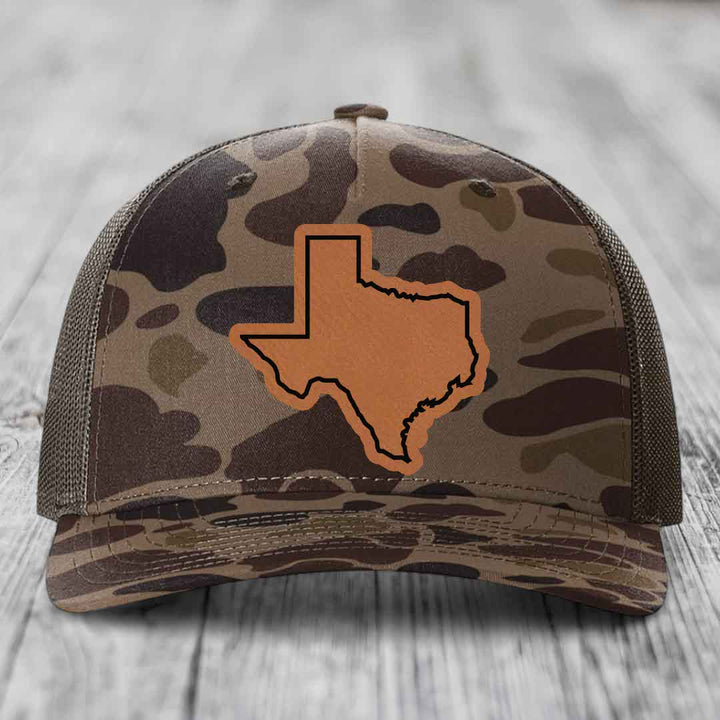 Camouflage trucker hat with brown Texas state outline patch on front.
