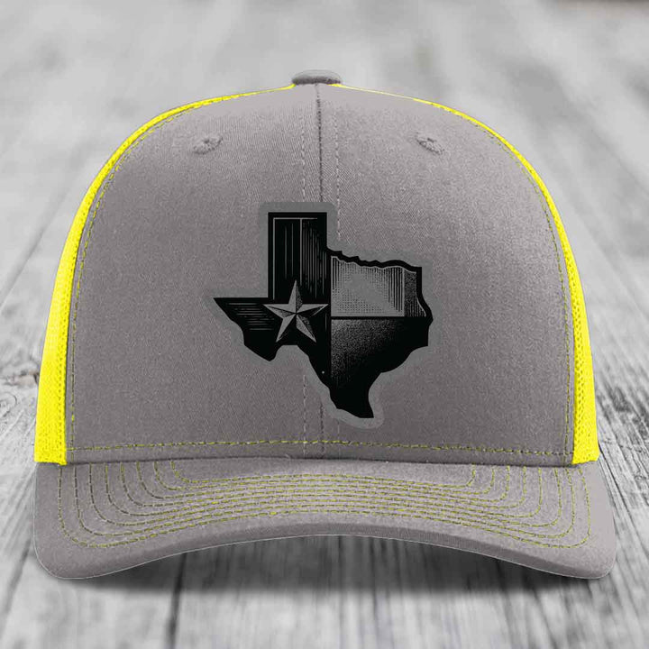 Grey trucker hat with black Texas-shaped patch featuring star and stripes, yellow mesh back and stitching.