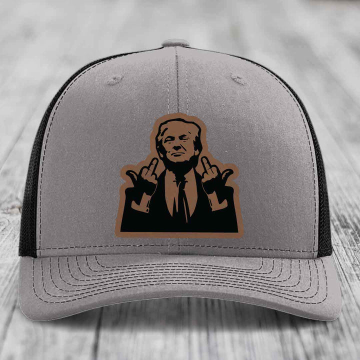 Grey trucker hat with black mesh back and brown leather patch of a man giving two middle fingers.