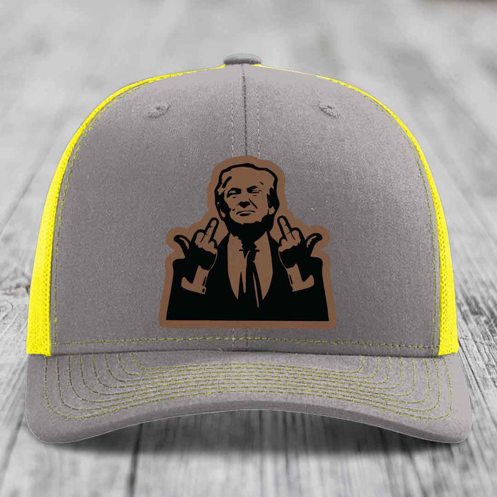 Grey trucker hat with yellow mesh sides and stitching, featuring a brown leatherette patch of a man making hand gestures.