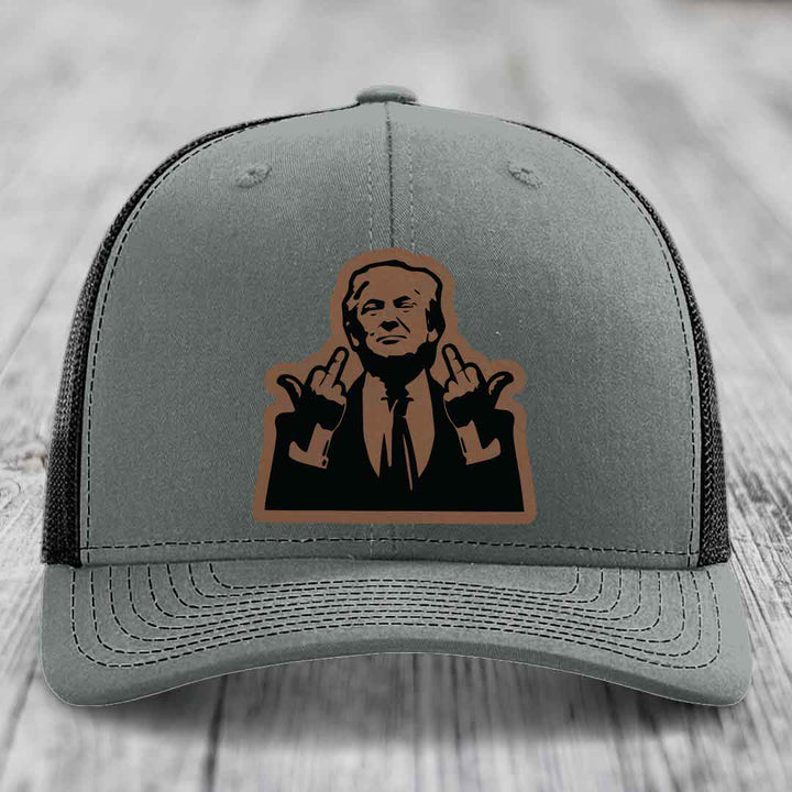 Grey trucker hat with black mesh back and brown leather patch of man giving middle finger