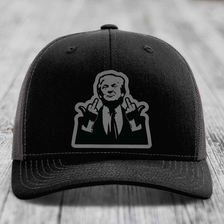 Black trucker hat with gray graphic of man making hand gestures, on wooden surface.