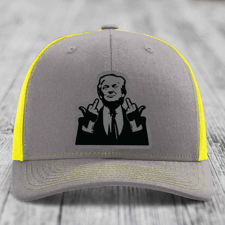 Grey trucker hat with yellow mesh and stitching, featuring a black patch of a man making rude hand gestures.