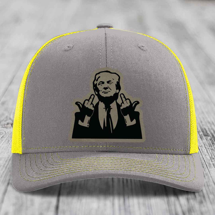 Grey trucker hat with yellow mesh and stitching, featuring a black patch of a man making rude hand gestures.