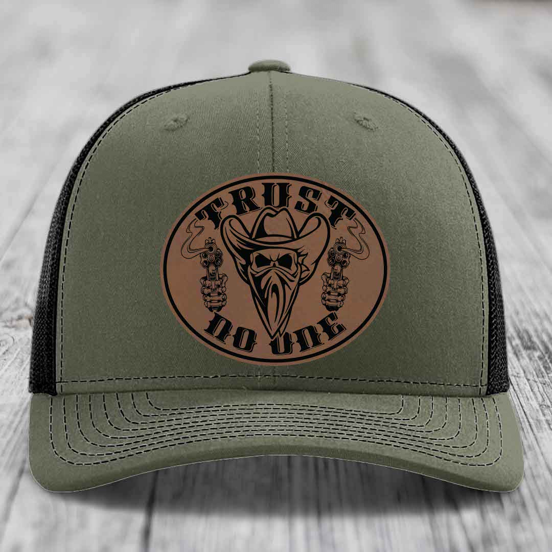 Olive green trucker hat with brown leather patch featuring skull, cowboy hat, and guns with &