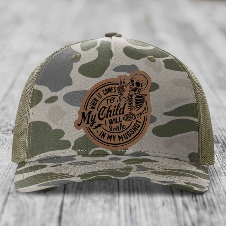 Camouflage trucker hat with a brown leather patch featuring a skeleton holding peace sign and text &