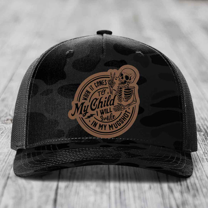 Black camouflage trucker hat with a brown leather patch featuring a skeleton holding up two fingers and text reading &