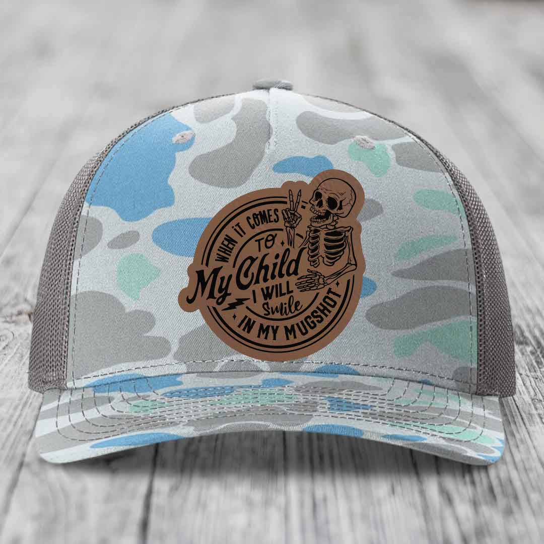 Camouflage trucker hat with brown leather patch featuring skeleton holding peace sign and text &
