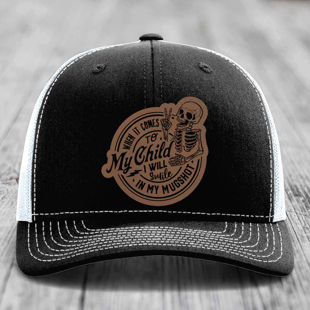 Black trucker hat with white mesh back and stitching, featuring a brown leather patch of a skeleton making a peace sign with text &