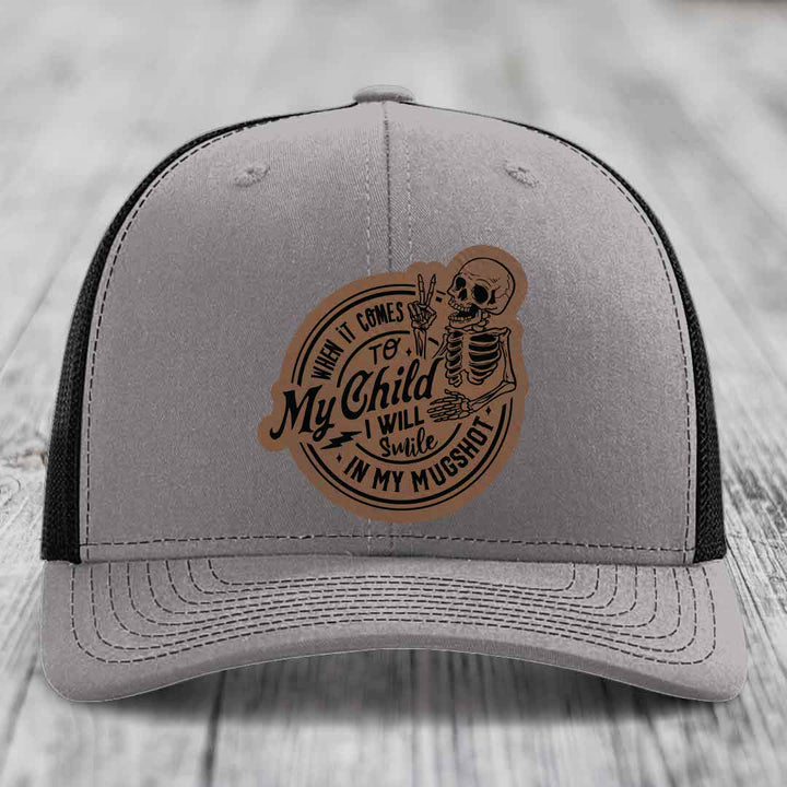 Grey trucker hat with black mesh back and a brown leatherette patch featuring a skeleton holding a peace sign and the text &