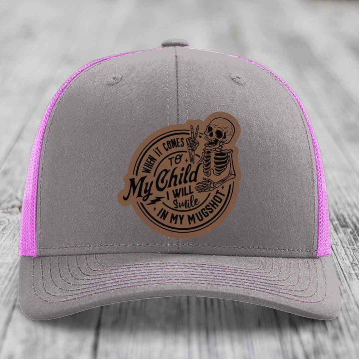 Grey trucker hat with pink mesh and a brown leatherette patch of a skeleton holding up peace sign with text &