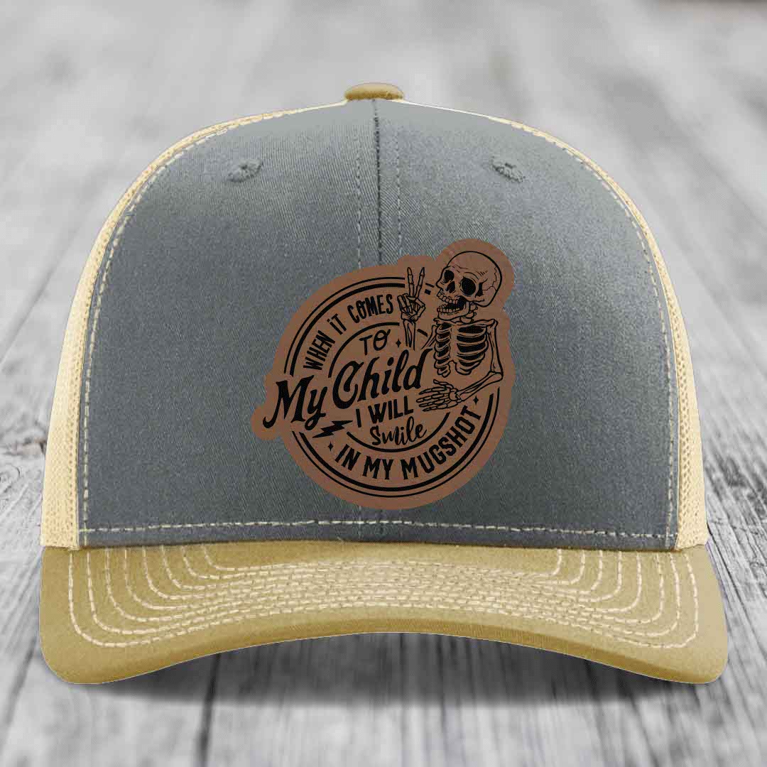 Grey and beige trucker hat with a brown leather patch featuring a skeleton holding up two fingers and the text &