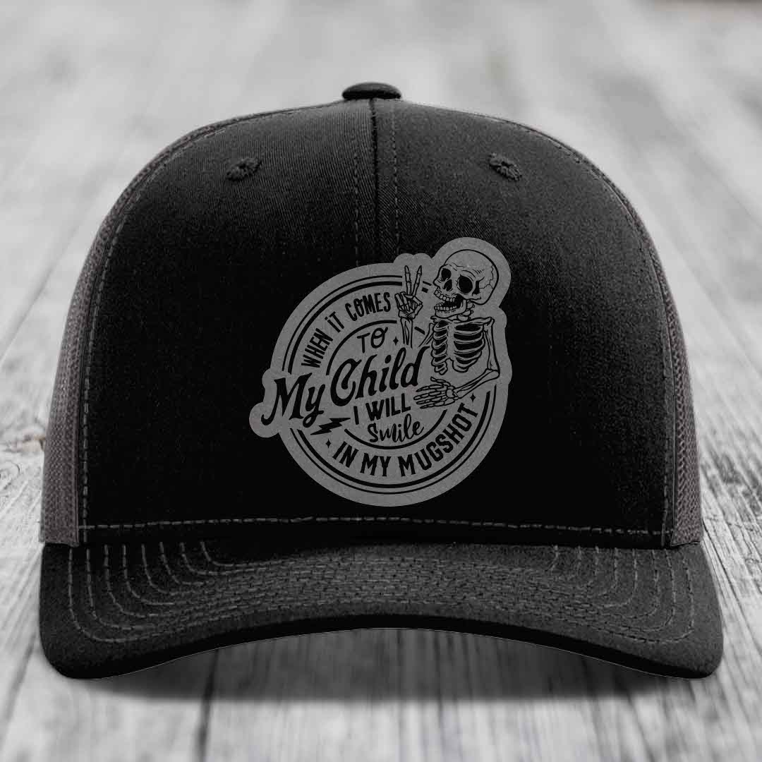Black trucker hat with a grey circular patch featuring a skeleton holding a peace sign and text reading &