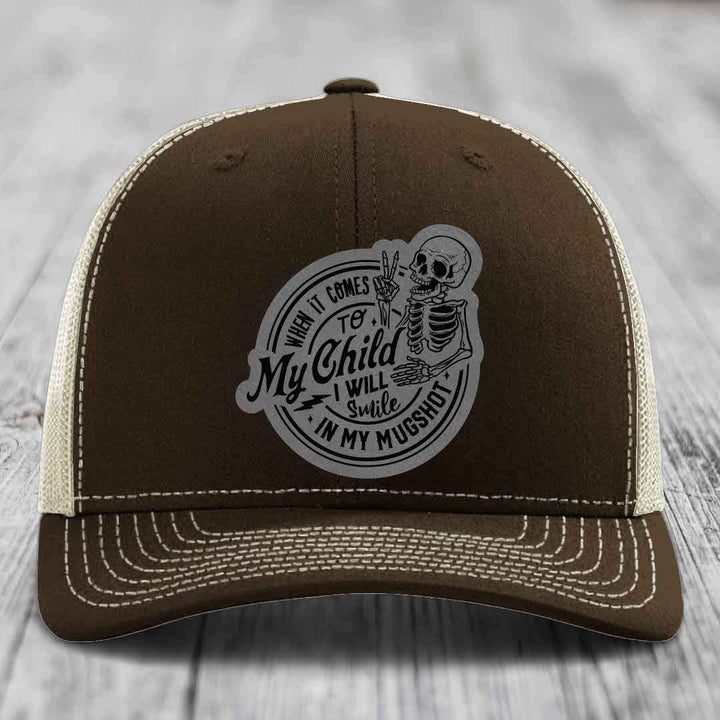 Brown trucker hat with white stitching and a grey circular patch featuring a skeleton holding up two fingers and the text &