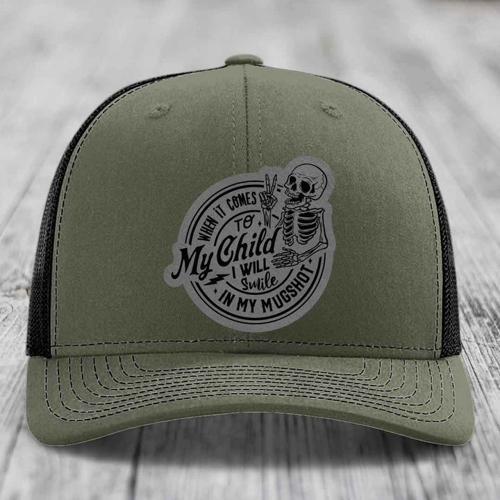 Olive green trucker hat with black mesh back and a circular patch featuring a skeleton making a peace sign.