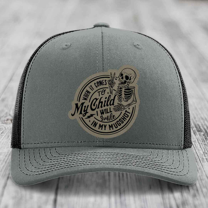Grey trucker hat with black mesh back and a circular patch featuring a skeleton making a peace sign and the text &