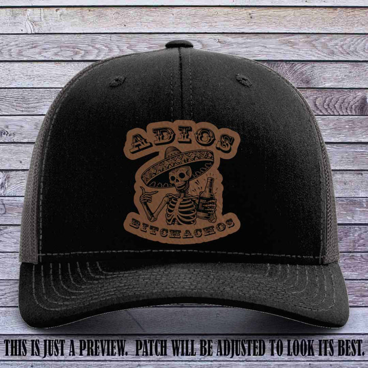 Dark brown leather patch on Black to Charcoal Richardson 112 trucker hat featuring “Adios Bitchachos” skeleton design.