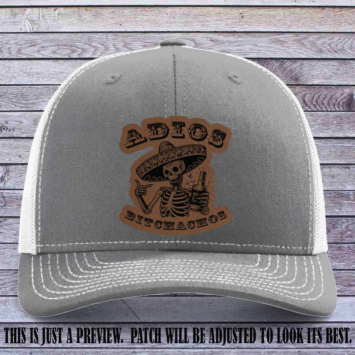 Dark brown leather patch on Heather Grey to White Richardson 112 trucker hat featuring “Adios Bitchachos” skeleton design.