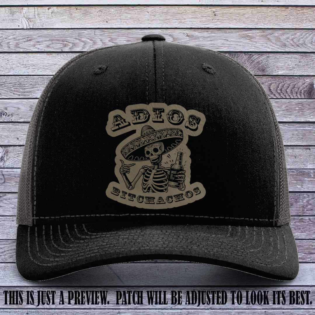 Light brown leather patch on Black to Charcoal Richardson 112 trucker hat featuring “Adios Bitchachos” skeleton design.