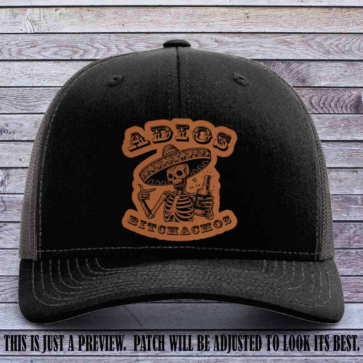 Rawhide leather patch on Black to Charcoal Richardson 112 trucker hat featuring “Adios Bitchachos” skeleton design.